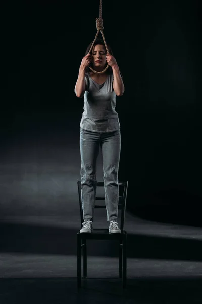 Mujer joven deprimida suicidándose mientras está de pie en la silla y poniendo soga en el cuello sobre fondo negro - foto de stock