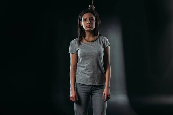 Depressive junge Frau blickt in die Kamera, während sie mit Schlinge am Hals vor schwarzem Hintergrund steht — Stockfoto