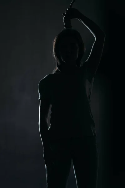 Депрессивная женщина совершает самоубийство стоя с петлей на шее в темноте на черном фоне — стоковое фото