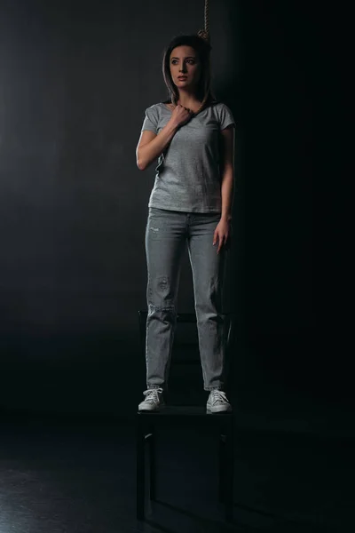 Depressed woman standing on chair with noose on neck and looking away on black background — Stock Photo