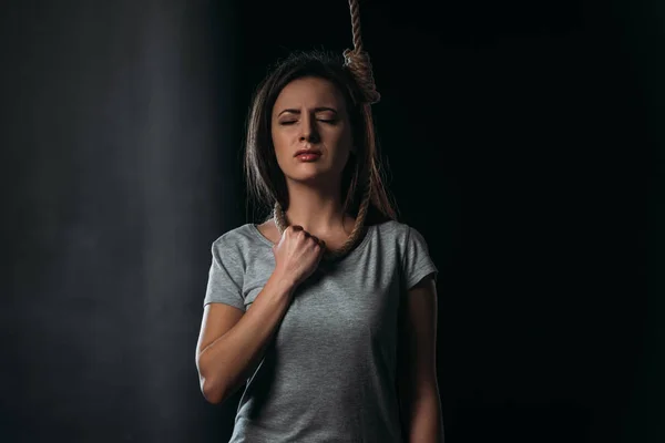 Безнадежная женщина совершает самоубийство стоя с петлей на шее на черном фоне — стоковое фото