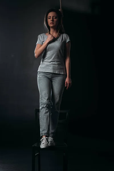 Занепокоєна жінка, яка здійснює самогубство, стоячи на стільці з носом на шиї на чорному тлі — стокове фото