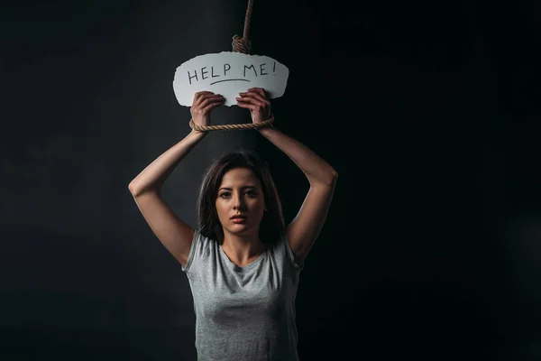 Frustrated young woman holding paper with help me inscription while standing under hanging noose and looking at camera on black background — Stock Photo