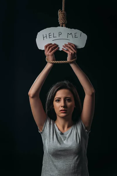Frustrierte junge Frau hält Papier mit Hilfe von Schriftzügen in der Hand, während sie unter hängender Schlinge steht und isoliert auf schwarz in die Kamera schaut — Stockfoto