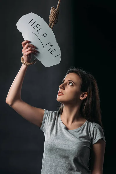 Mujer joven deprimida sosteniendo papel con ayuda me letras cerca de colgar la soga en el fondo negro - foto de stock