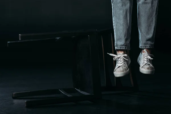 Cropped view of self-murder legs hanging near fallen chair on black background — Stock Photo