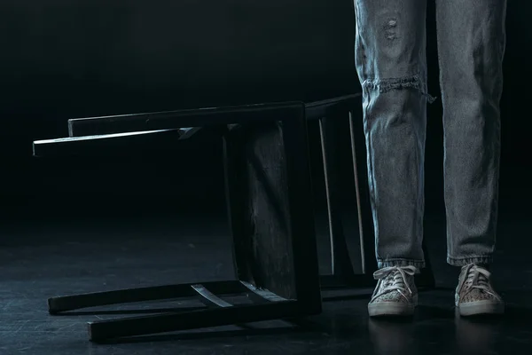 Vista recortada de la mujer de pie cerca de la silla caída sobre fondo negro - foto de stock