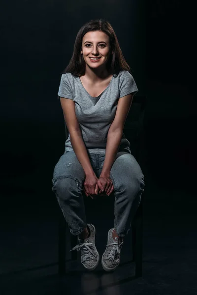 Glückliche junge Frau lächelt in die Kamera, während sie im Dunkeln auf schwarzem Hintergrund sitzt — Stockfoto