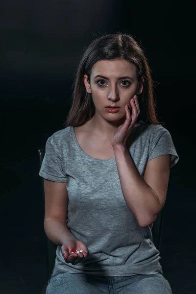 Mujer deprimida mirando a la cámara mientras sostiene un puñado de pastillas y va a suicidarse aislado en negro - foto de stock