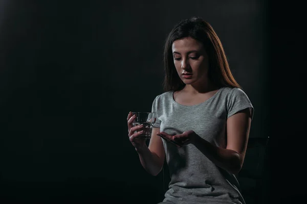 Депресивна жінка збирається покінчити життя самогубством, тримаючи жменьку таблеток і склянку води ізольовано на чорному — стокове фото