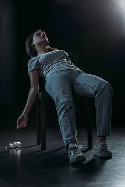 Mulher sem vida na cadeira cometeu suicídio por overdose pílulas no fundo escuro com iluminação — Fotografia de Stock