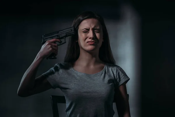 Llorando, mujer desesperada sosteniendo arma cerca de la barbilla sobre fondo oscuro - foto de stock