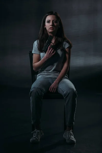 Verzweifelte Frau mit Pistole am Kinn und Blick in die Kamera, während sie auf Stuhl auf dunklem Hintergrund sitzt — Stockfoto