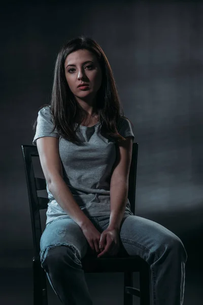 Frustrierte Frau blickt in die Kamera, während sie in der Dunkelheit auf schwarzem Hintergrund auf einem Stuhl sitzt — Stockfoto