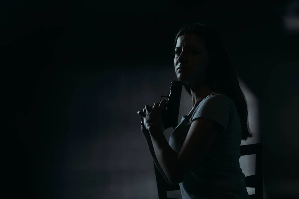 Расстроенная женщина смотрит в камеру, держа пистолет возле подбородка на темном фоне — стоковое фото