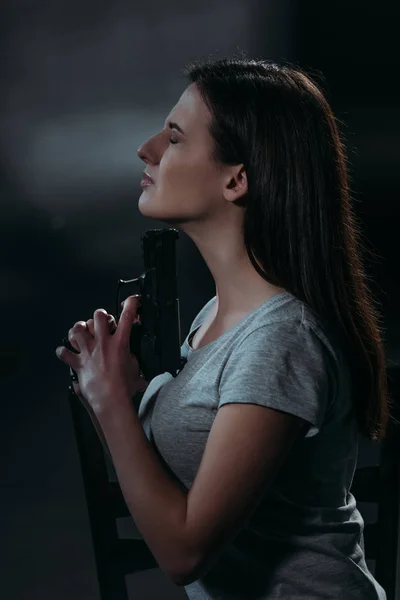 Vue latérale de la femme déprimée avec les yeux fermés tenant pistolet près du menton sur fond sombre — Photo de stock