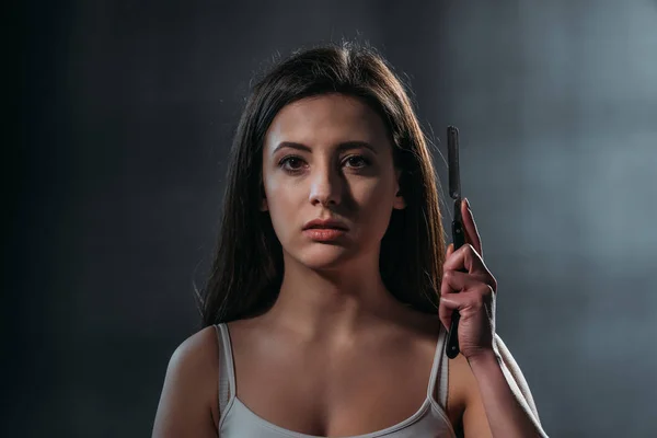 Depressive junge Frau blickt in Kamera und hält Rasiermesser auf dunklem Hintergrund — Stockfoto