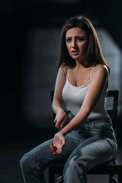 Mujer joven desesperada mirando a la cámara mientras se suicida cortando venas con cuchillo sobre fondo oscuro - foto de stock