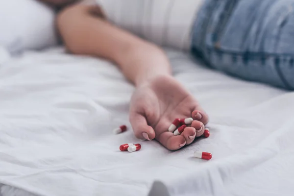 Partial view of lifeless woman, committed suicide by overdosing medicines, lying on bed near scattered pills — Stock Photo