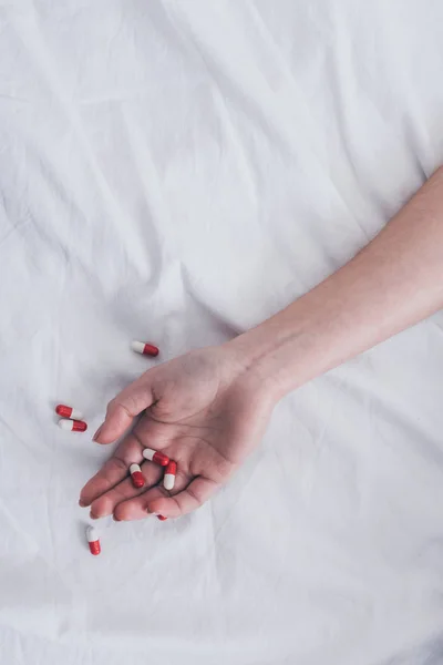 Vista cortada da mão feminina com pílulas na cama branca — Fotografia de Stock