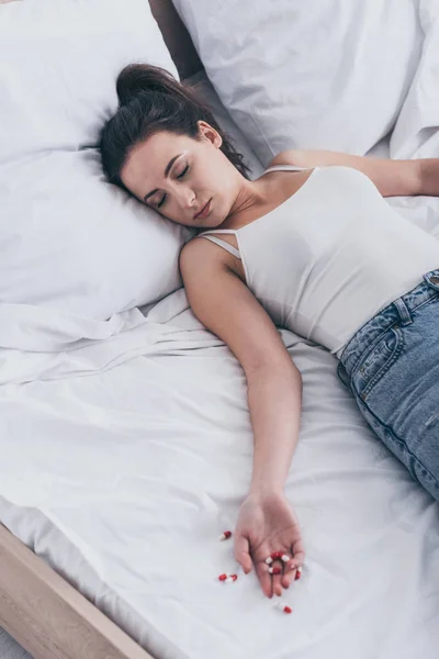 Частичный взгляд на безжизненную женщину, покончившую с собой передозировкой лекарств, лежащую на кровати возле разбросанных таблеток — стоковое фото