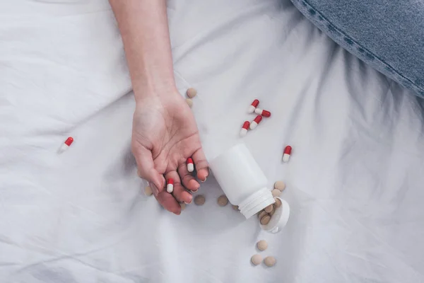 Partial view of lifeless woman, committed suicide by overdosing medicines, lying on bed near container with pills — Stock Photo