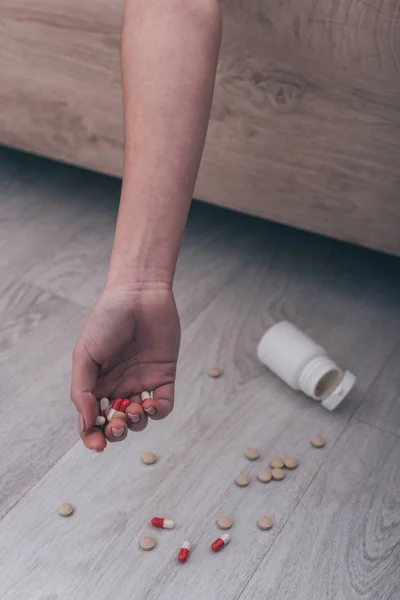 Обрезанный вид безжизненной женщины, покончившей с собой передозировкой лекарств, лежащей на кровати возле разбросанных таблеток на полу — стоковое фото