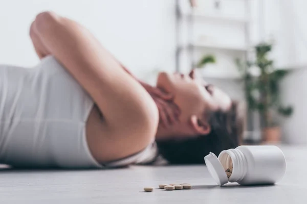 Selective focus of dying woman, committing suicide by overdosing medicines, lying on floor near container with pills and touching throat — Stock Photo