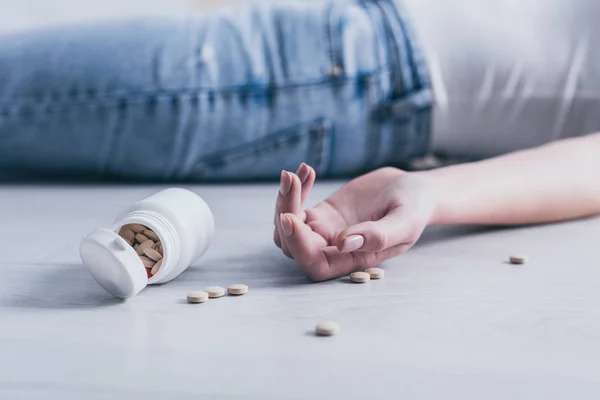 Partial view of lifeless woman, committed suicide by overdosing medicines, lying on floor near container with pills — Stock Photo