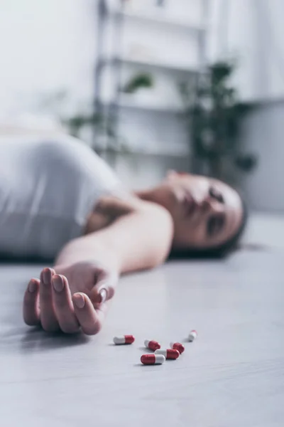 Selektiver Fokus des leblosen Mädchens, das Selbstmord durch Überdosierung von Medikamenten beging, auf dem Boden liegend in der Nähe verstreuter Tabletten — Stockfoto
