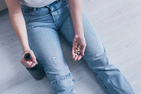 Vista ritagliata di donna seduta sul pavimento con bottiglia di alcol e una manciata di pillole — Foto stock