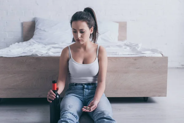 Разочарованная молодая женщина сидит на полу с бутылкой алкоголя и собирается покончить жизнь самоубийством, принимая лекарства — стоковое фото