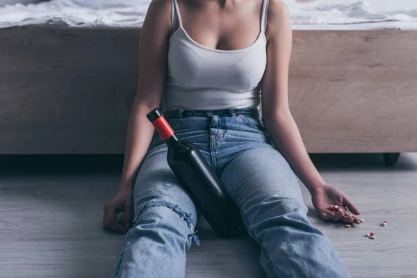 Частичный вид женщины, сидящей на полу с бутылкой алкоголя и горсткой таблеток — стоковое фото