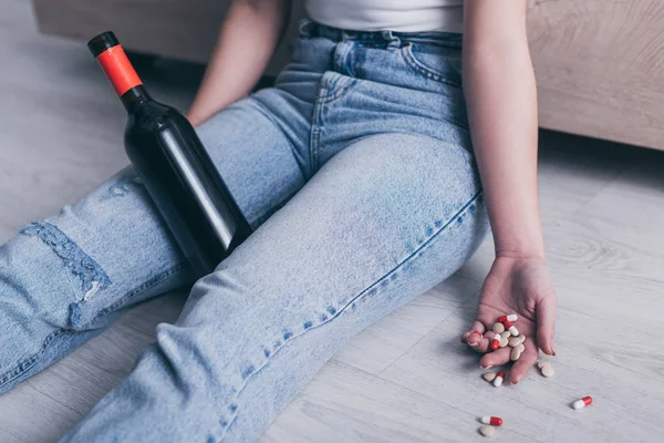 Vista ritagliata di donna incosciente seduta sul pavimento con bottiglia di alcol e una manciata di pillole — Foto stock