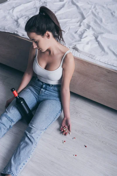 Mulher inconsciente sentado no chão com garrafa de álcool e punhado de comprimidos — Fotografia de Stock