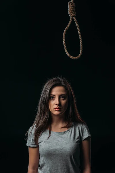 Depressed young woman looking away while standing under hanging noose isolated on balck — Stock Photo