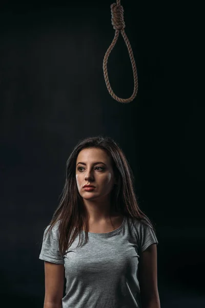 Frustrated young woman looking away while standing under hanging rope noose isolated on balck — Stock Photo