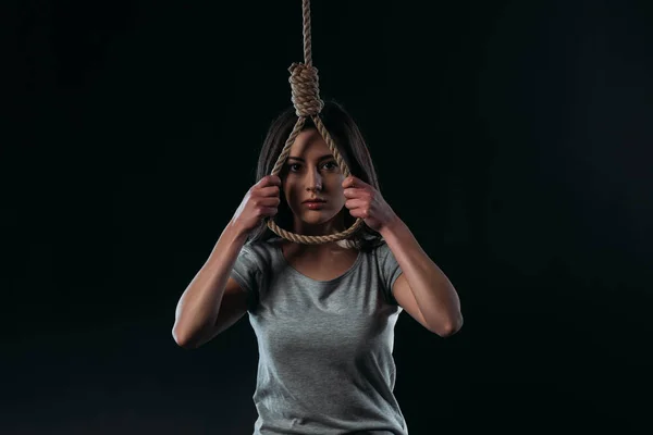 Пригнічена молода жінка збирається покінчити життя самогубством, тримаючи висячу мотузку і дивлячись на камеру ізольовано на чорному — стокове фото