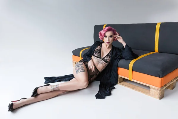Привлекательная девушка с татуировками в прозрачном нижнем белье и в плаще позирует на диване — стоковое фото