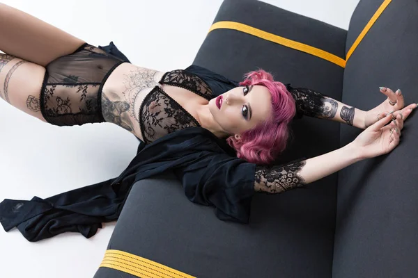 Seductora chica de pelo rosa tatuada en lencería transparente posando en el sofá - foto de stock