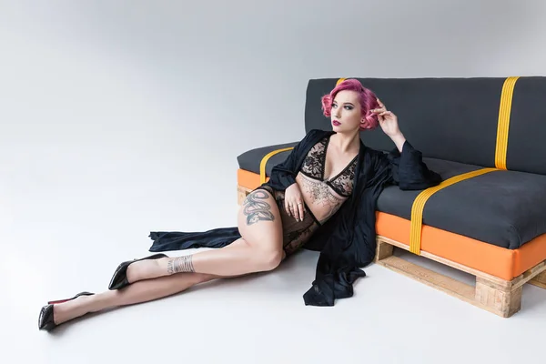 Hermosa tatuada pin up chica en lencería transparente posando en el sofá - foto de stock