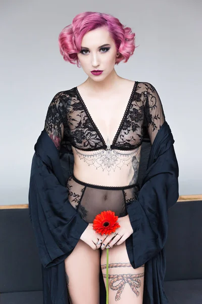 Seducente tatuato pin up ragazza in lingerie trasparente con fiore posa accanto al divano — Foto stock