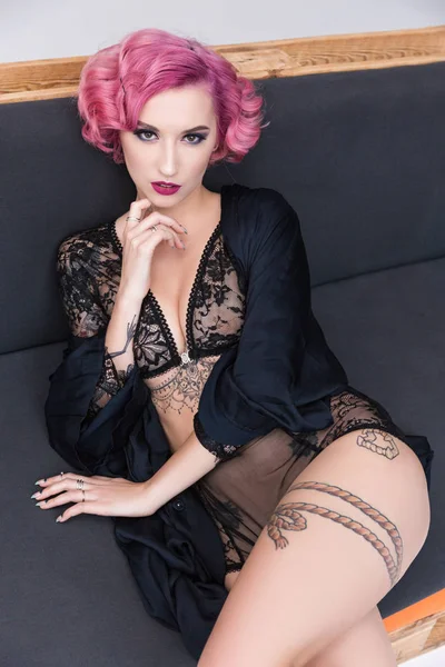Соблазнительная татуированная розововолосая девушка в прозрачном белье позирует на диване — стоковое фото