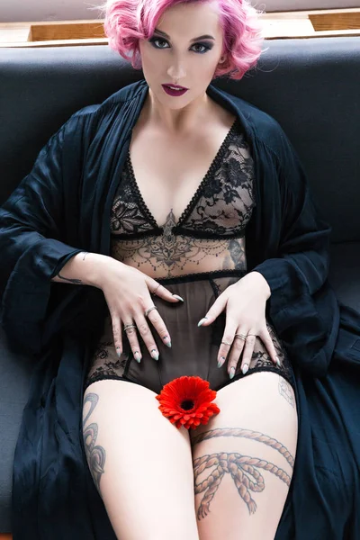 Seductora chica tatuada pin up en lencería transparente con flor posando en el sofá - foto de stock
