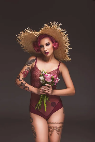 Atractiva chica tatuada pin up en lencería y sombrero de paja posando con flores delante de fondo gris - foto de stock