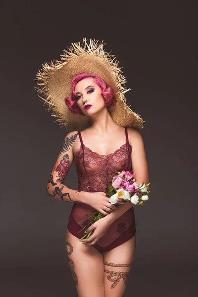 Graziosa ragazza tatuata in lingerie e cappello di paglia con fiori davanti a sfondo grigio — Foto stock