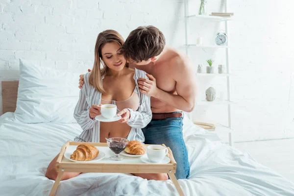 Homme torse nu embrassant femme avec tasse de café près du plateau sur la literie — Photo de stock